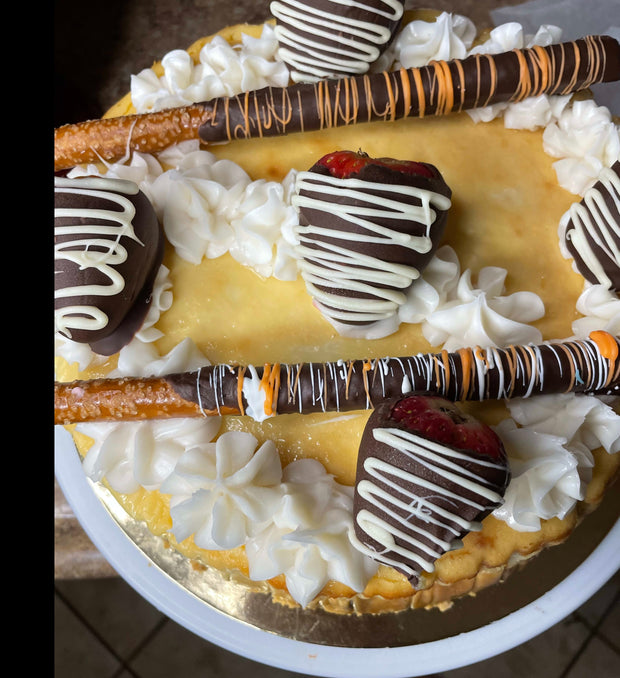 A dense, creamy New York Style Cheesecake Topped with custom chocolate pretzels, chocolate-covered strawberries, cream cheese icing, and a pretzel crust. A heavenly rich classic dessert.