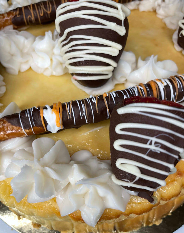 A dense, creamy New York Style Cheesecake Topped with custom chocolate pretzels, chocolate-covered strawberries, cream cheese icing, and a pretzel crust. A heavenly rich classic dessert.